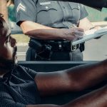 A police officer issues a traffic ticket to a driver seated in a car during the day.