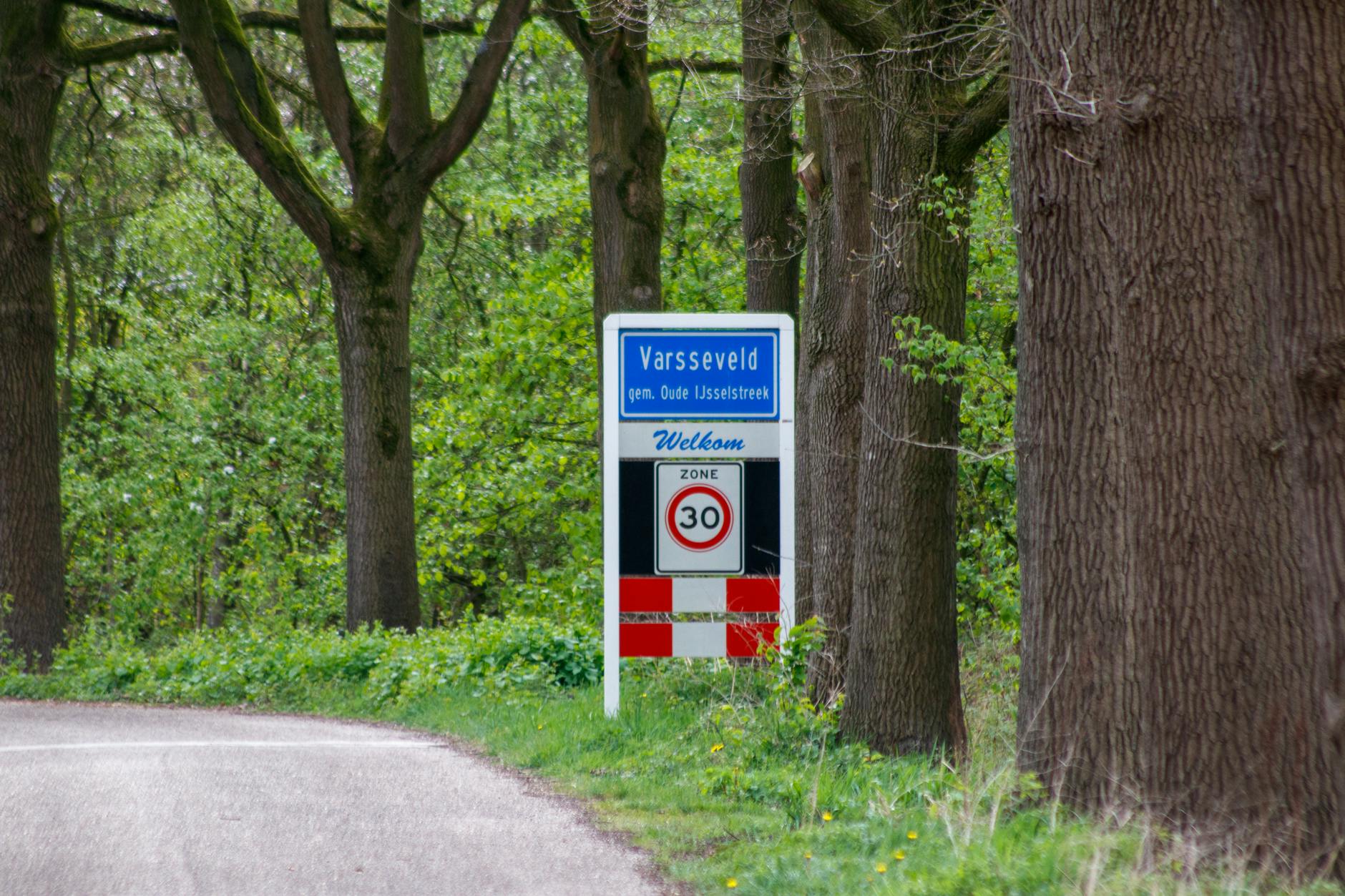 Sign welcoming to Varsselveld, nestled in a green, leafy roadside setting.