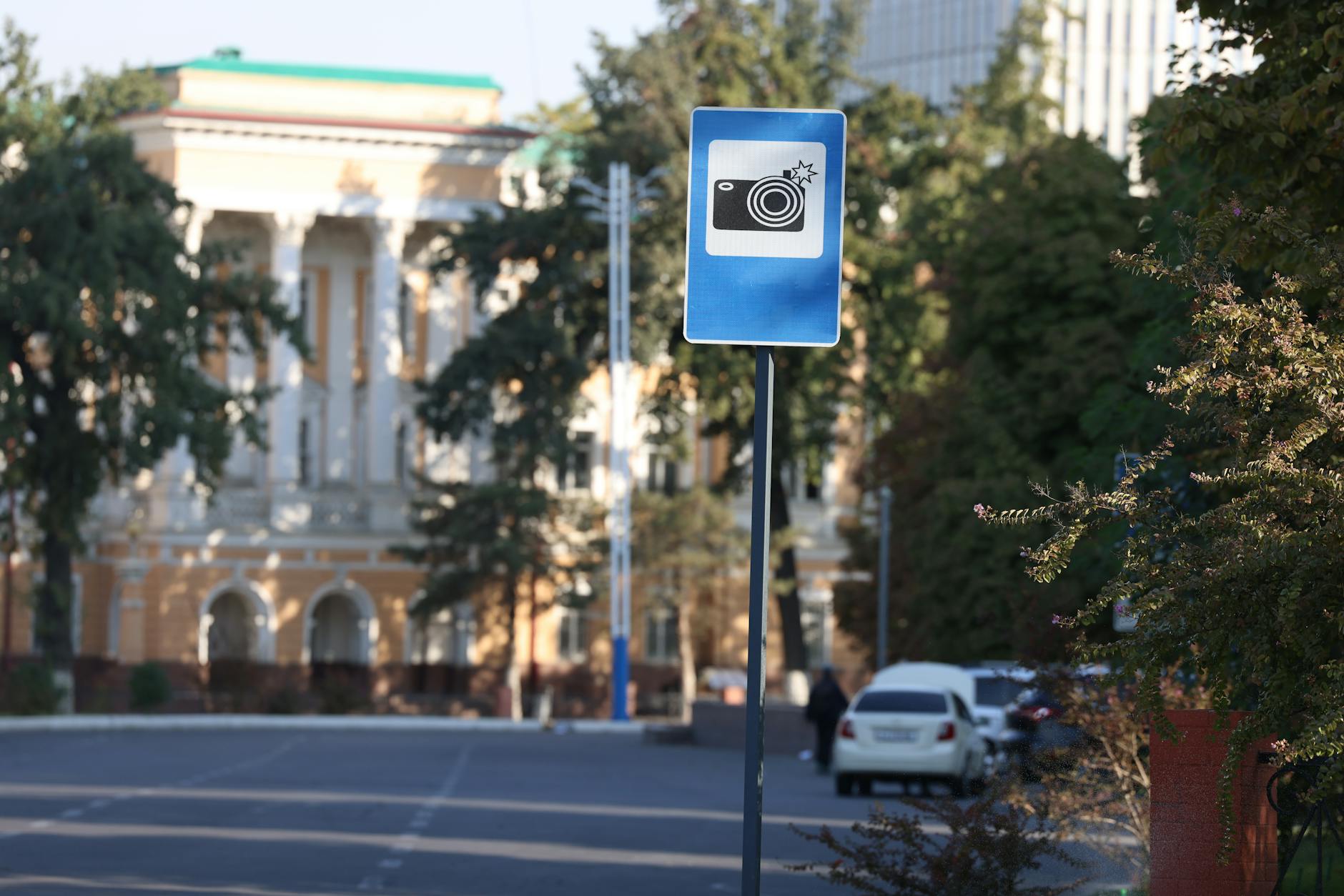 A blue traffic camera warning sign on a city street with blurred buildings in the background.