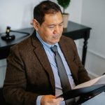From above of serious ethnic lawyer in elegant suit checking report in folder on blurred background of office