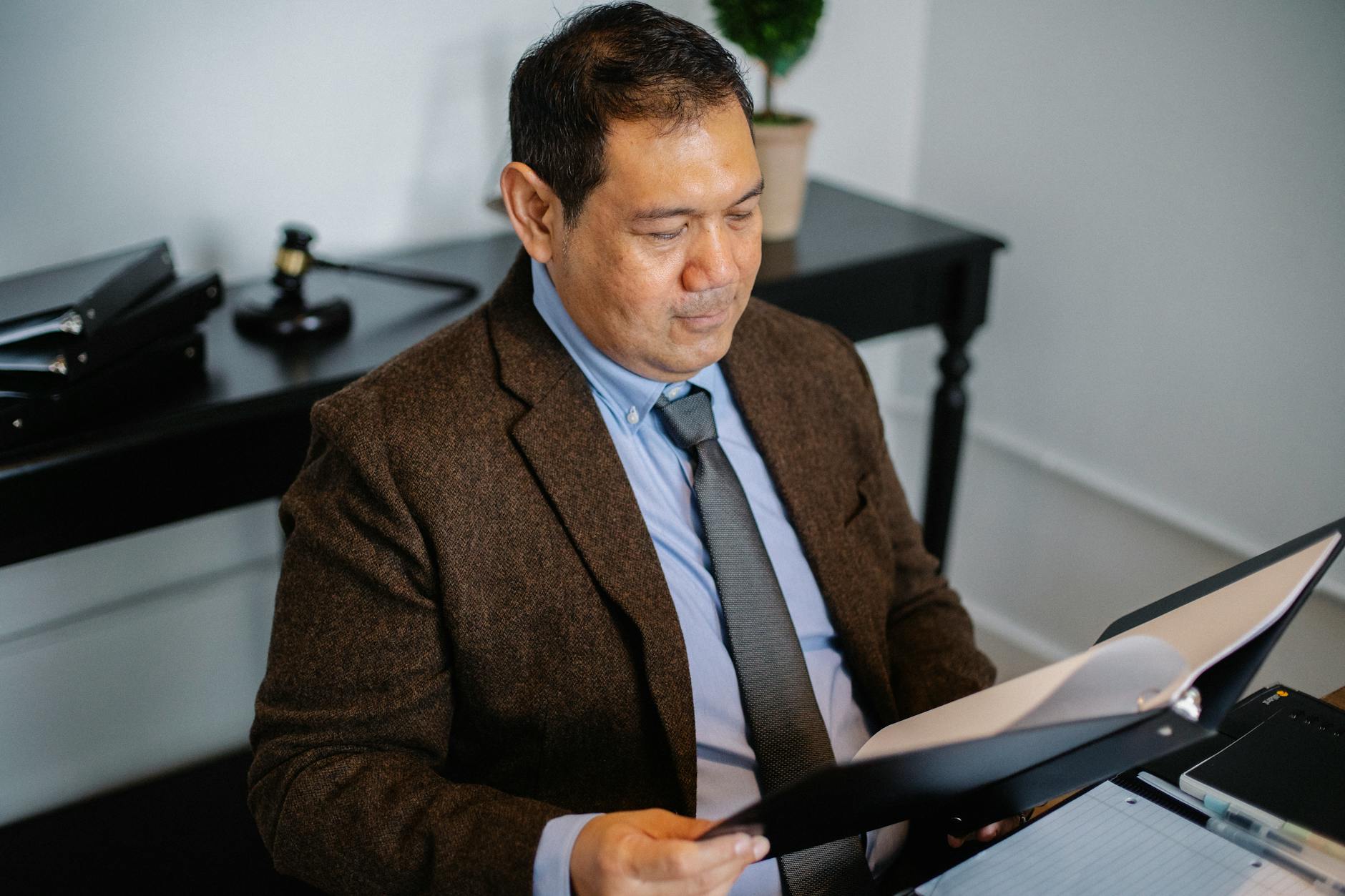 From above of serious ethnic lawyer in elegant suit checking report in folder on blurred background of office