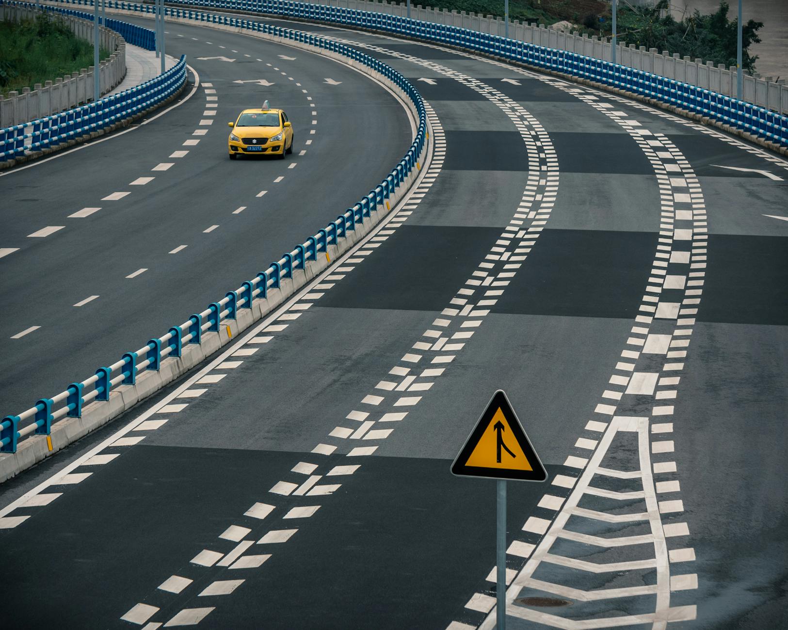 A lone yellow taxi navigates around a curved, empty highway with a merge sign visible.