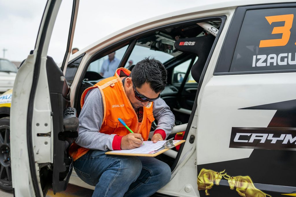 A motorsport official reviews documents while sitting by a rally car during a race event.