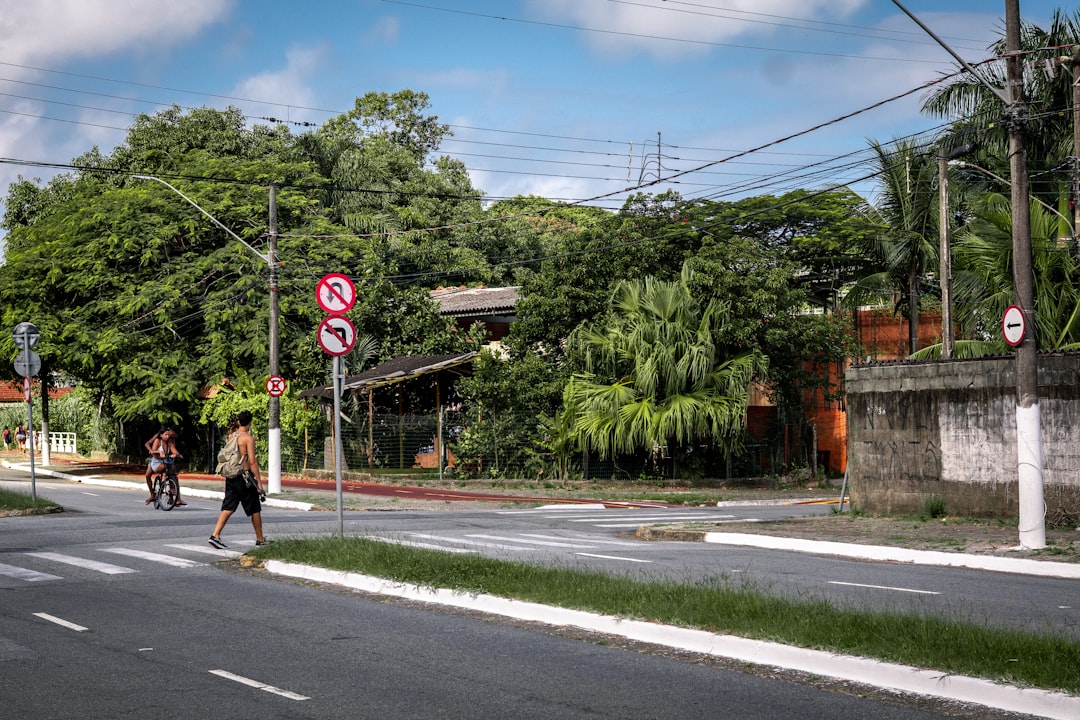 Homem Atravessa Rua Com Arvores Exuberantes Ao Fundo Ga1r0SYCoTg