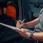 A police officer writes a ticket as the driver looks on from inside the car.