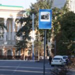 A blue traffic camera warning sign on a city street with blurred buildings in the background.