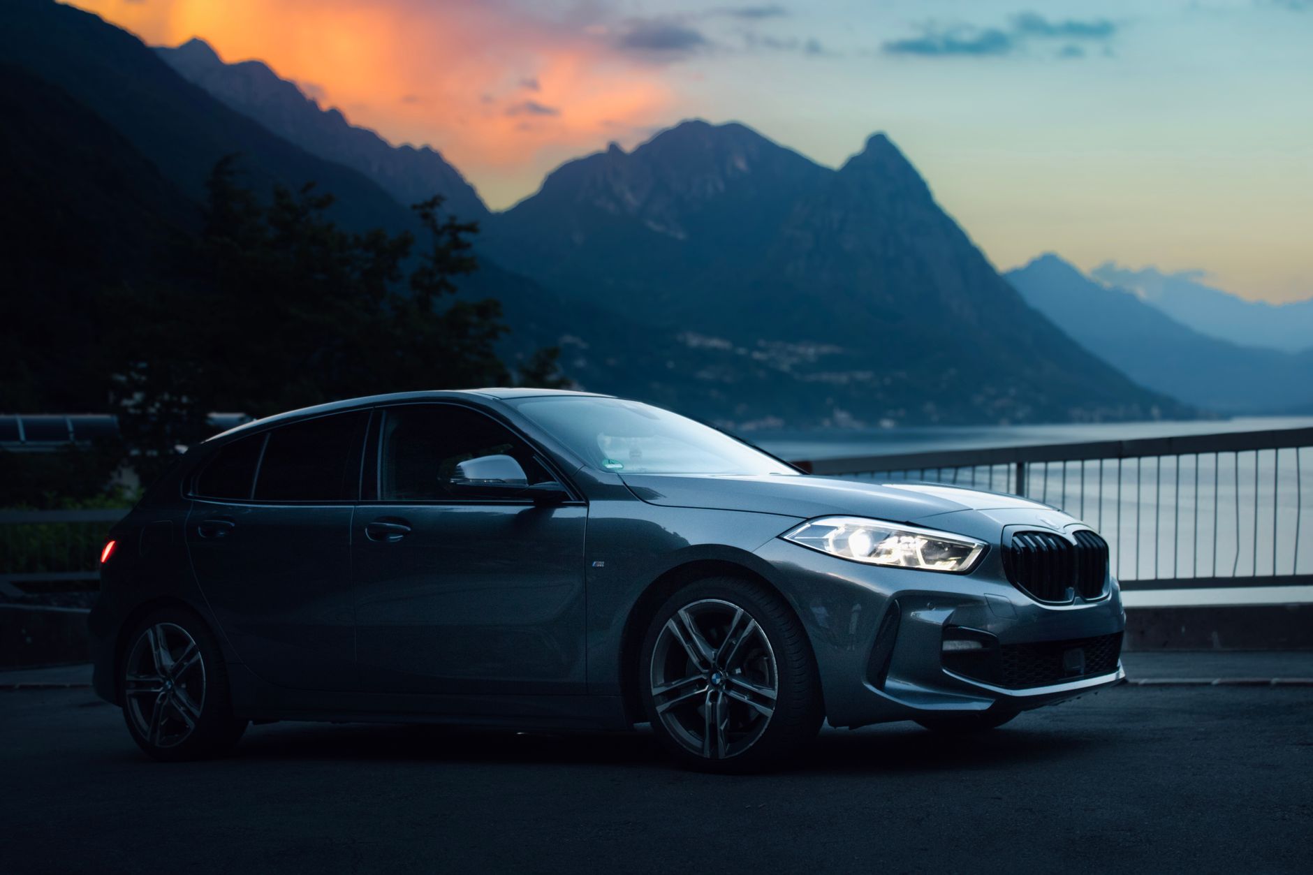 A sleek luxury car parked by Lake Como with breathtaking sunset and mountains in the backdrop.