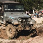 Jeep navigating a muddy track during an off-road event with spectators in the background.
