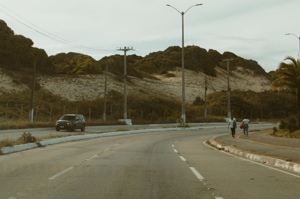 A peaceful road view in Natal, Brazil, featuring hilly terrain, a car, and pedestrians under a cloudy sky.