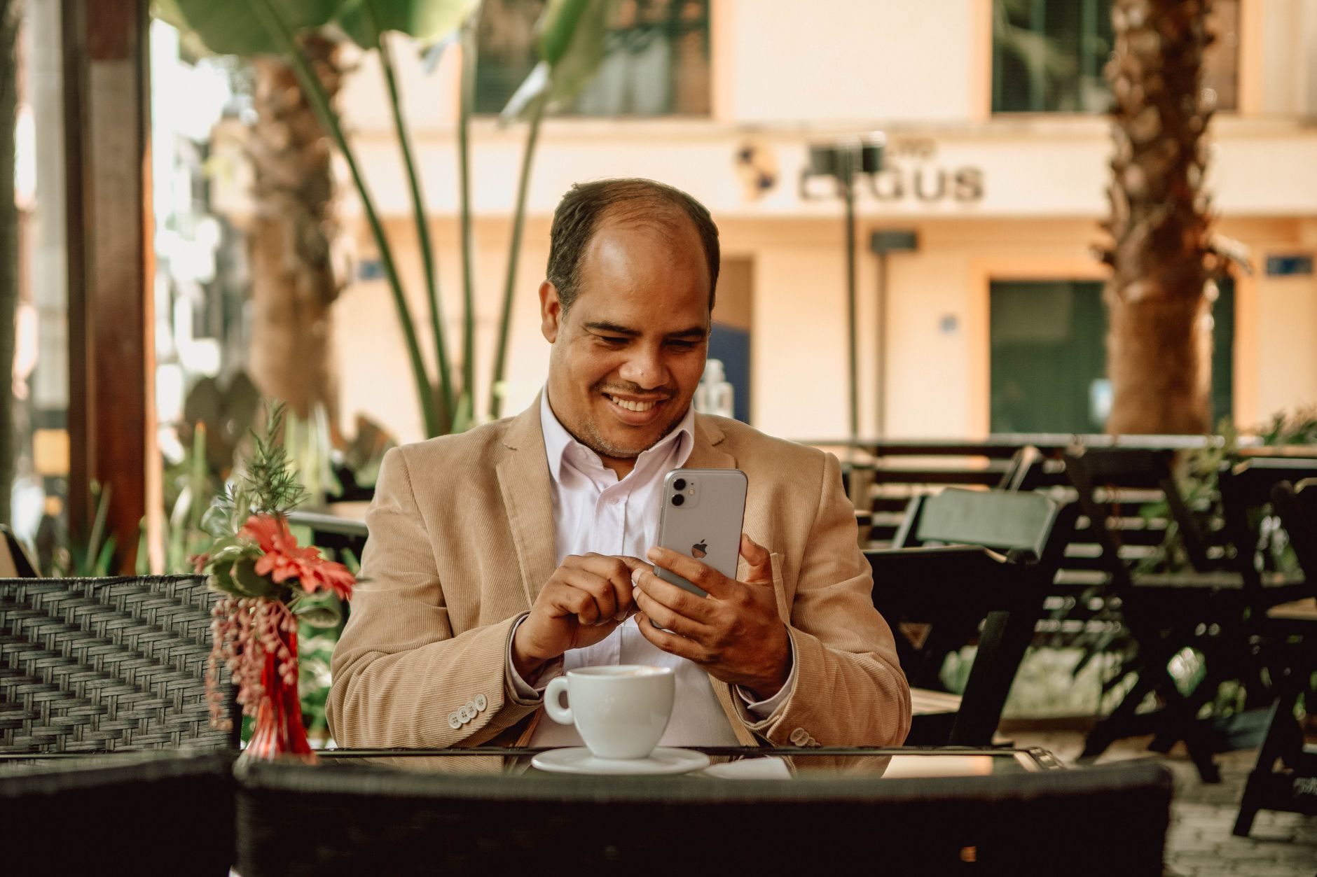A man in a white shirt smiles while using his smartphone at an outdoor café.