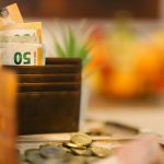 Brown wallet holding banknotes with scattered coins on a wooden table.