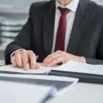 Close-up of a businessman reviewing documents at an office desk. Focus on hands and paperwork.