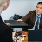 Serious middle aged ethnic male manager in suit giving document folder to unrecognizable female colleague during work at table with laptop in office