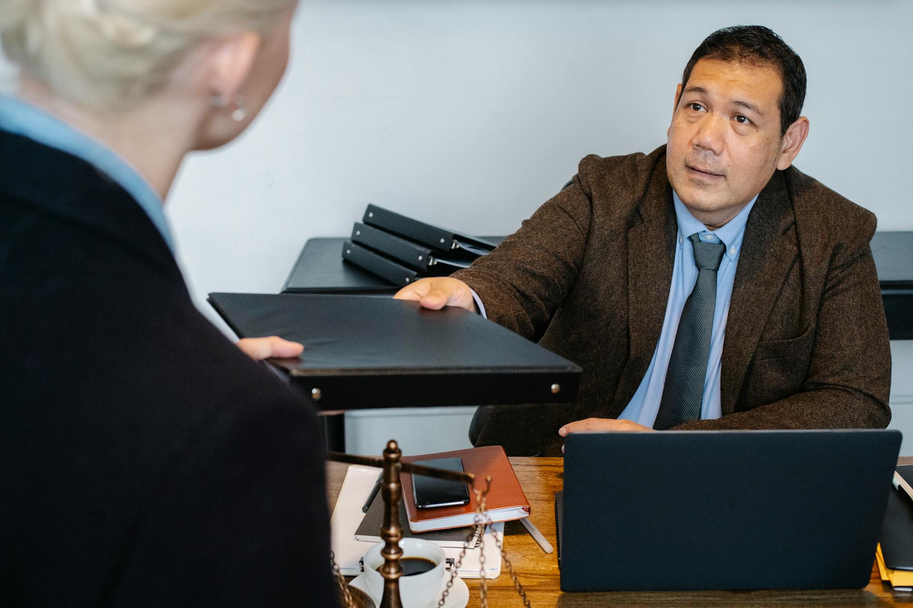 Serious middle aged ethnic male manager in suit giving document folder to unrecognizable female colleague during work at table with laptop in office