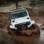 A white jeep navigates through a challenging muddy off-road track during a competition.