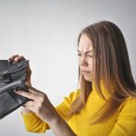 Young woman in a yellow top looking at her empty wallet with a frustrated expression.