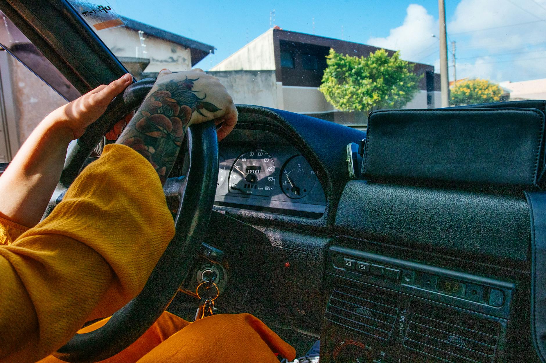 Close-up of a tattooed person driving a vintage car with a colorful dashboard.