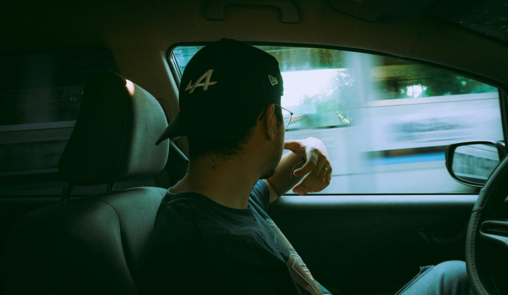 Rear view of a man driving a car, wearing a baseball cap and glasses in daylight.