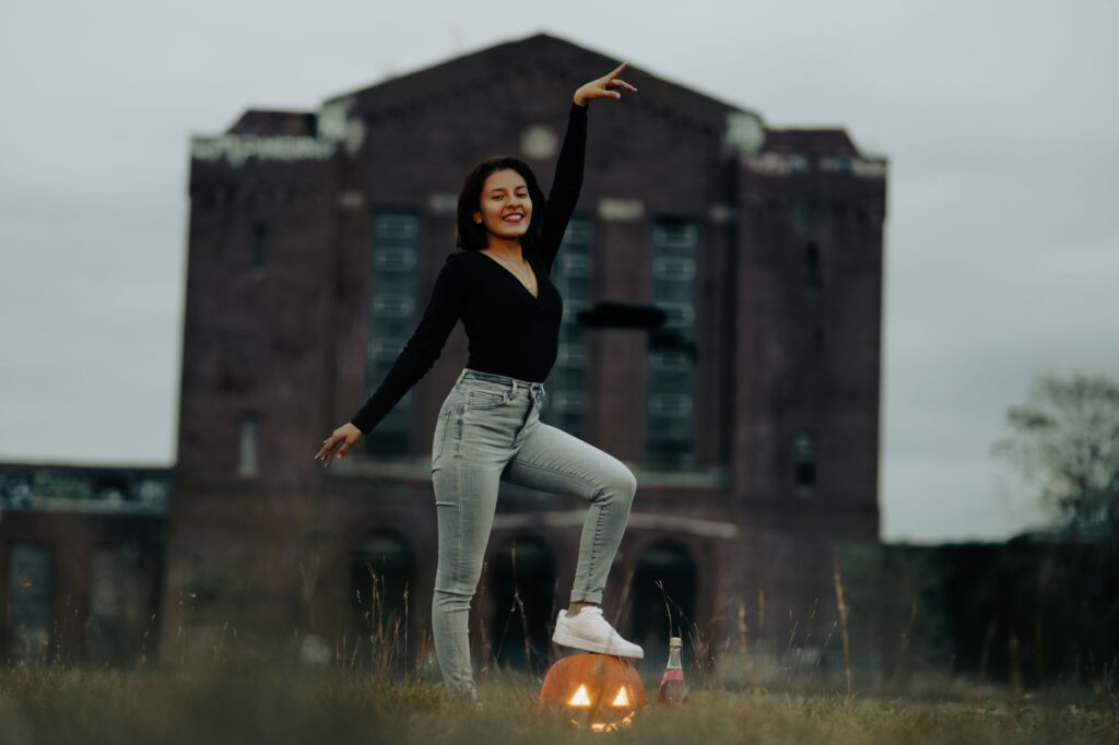A woman poses with a carved pumpkin in front of an abandoned building, celebrating Halloween.