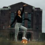 A woman poses with a carved pumpkin in front of an abandoned building, celebrating Halloween.