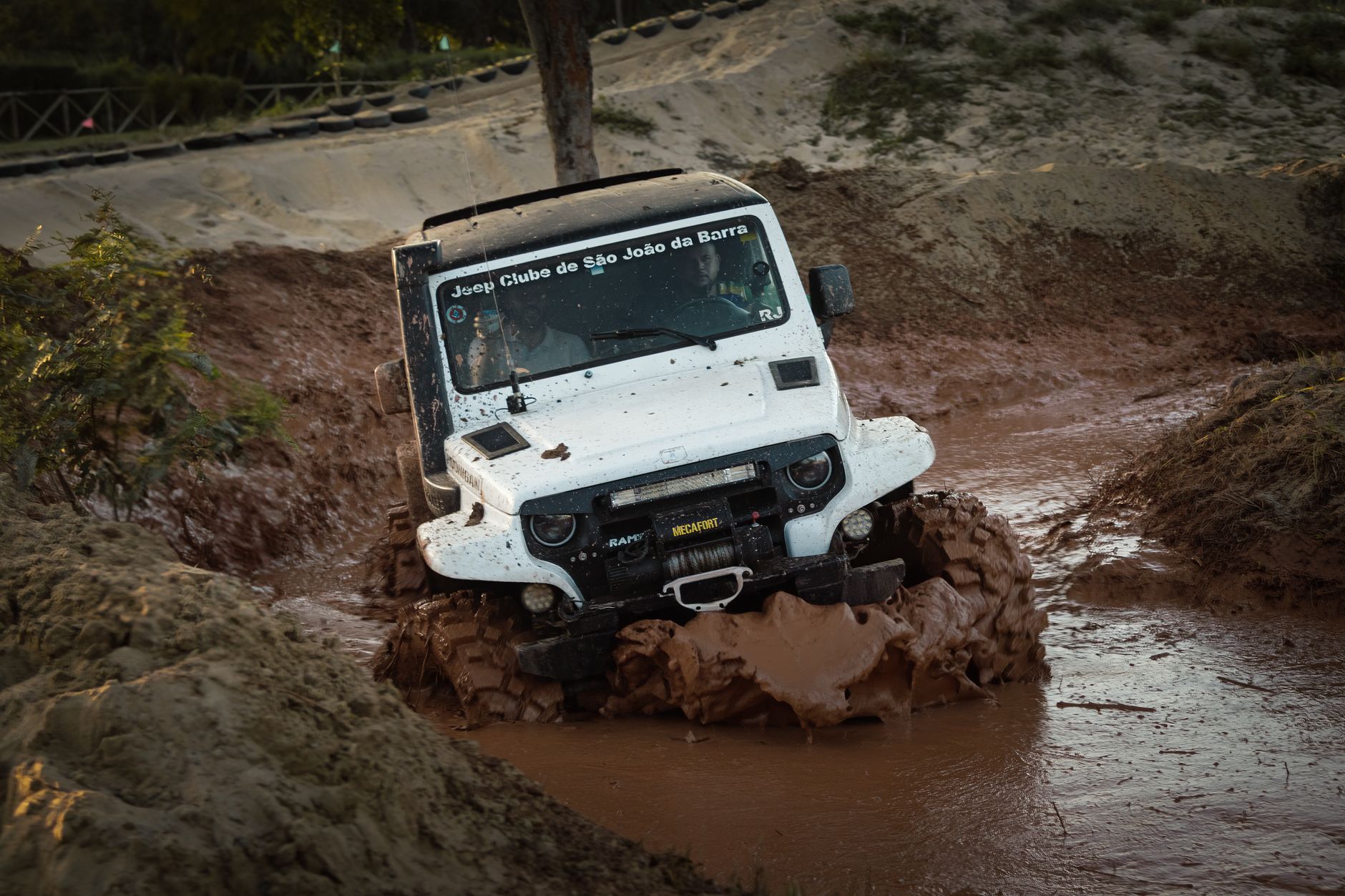 A white jeep navigates through a challenging muddy off-road track during a competition.