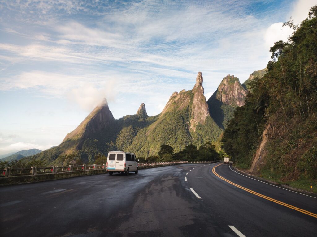 Van traveling on a scenic road towards the iconic Dedo de Deus mountain under a bright sky.
