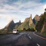 Van traveling on a scenic road towards the iconic Dedo de Deus mountain under a bright sky.