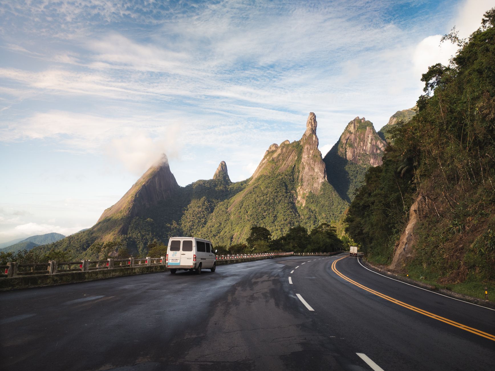 Van traveling on a scenic road towards the iconic Dedo de Deus mountain under a bright sky.