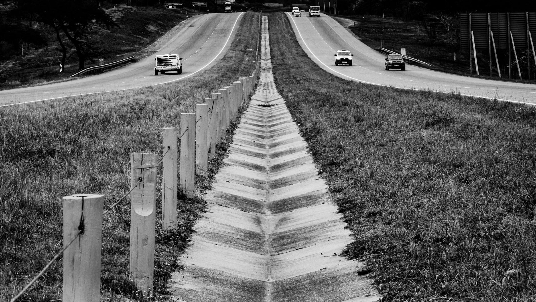 Monochrome view of a highway with cars and a wooden fence in the foreground.