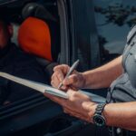 A police officer writes a ticket as the driver looks on from inside the car.