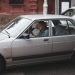 A classic silver sedan with a driver using a mobile phone in urban traffic.