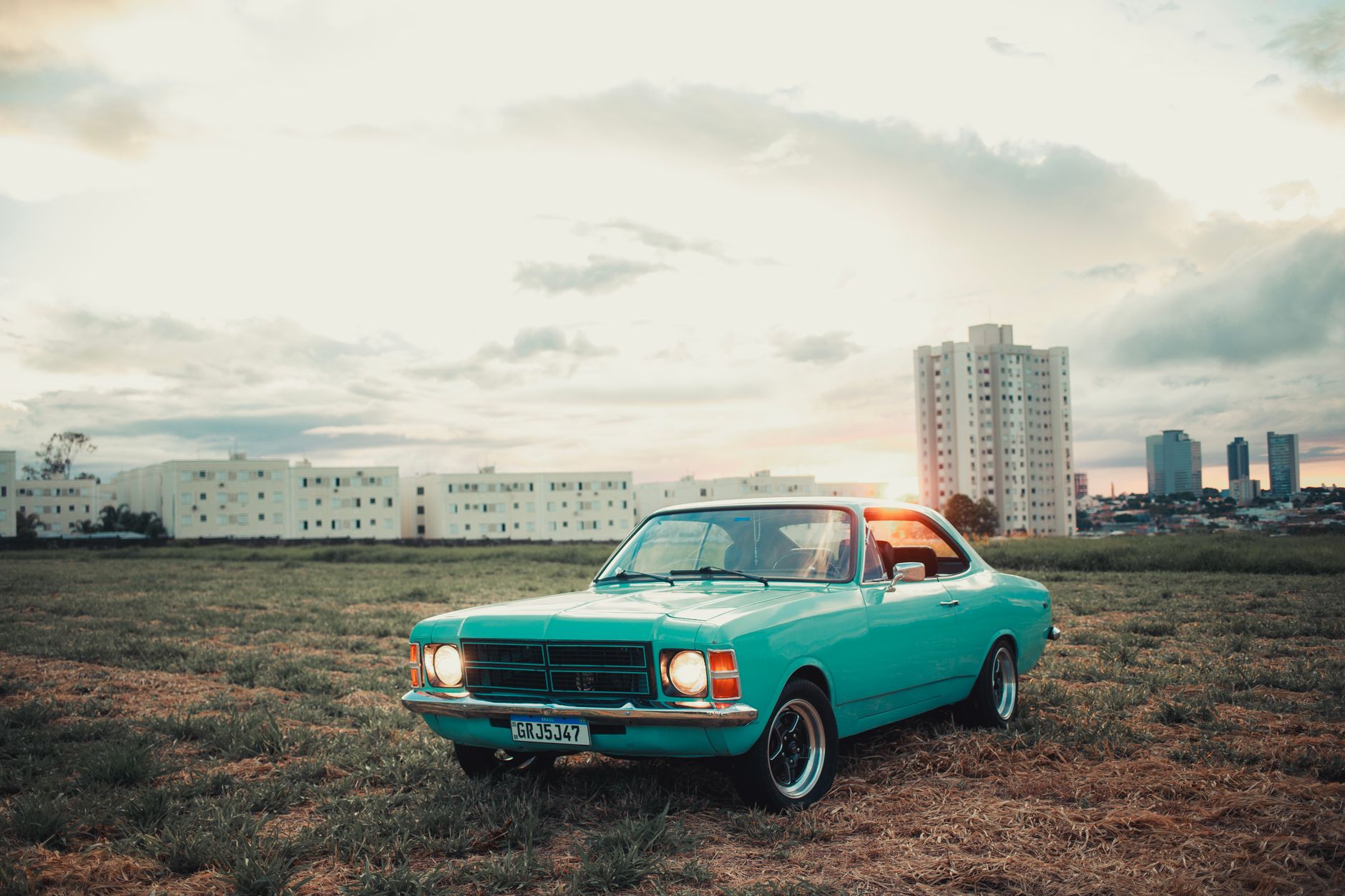 A vintage turquoise car parked in a field with a city skyline backdrop at sunset, capturing urban nostalgia.