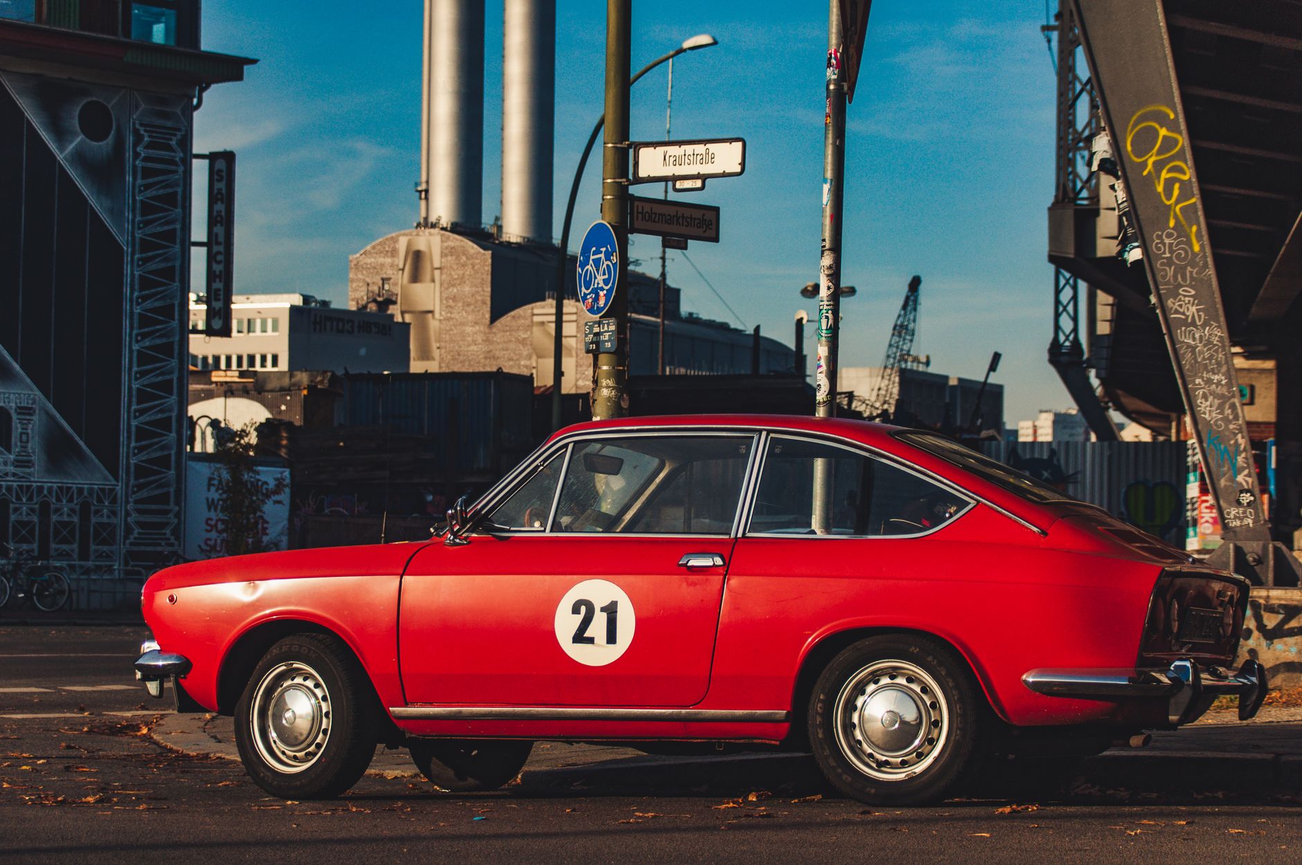 A classic red car with a retro design parked on an industrial city street under a bright blue sky.