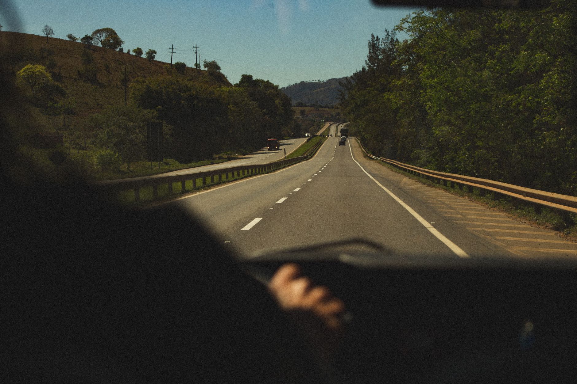 View from inside transport of unrecognizable traveler driving on asphalt roadway between lush green trees under blue sky