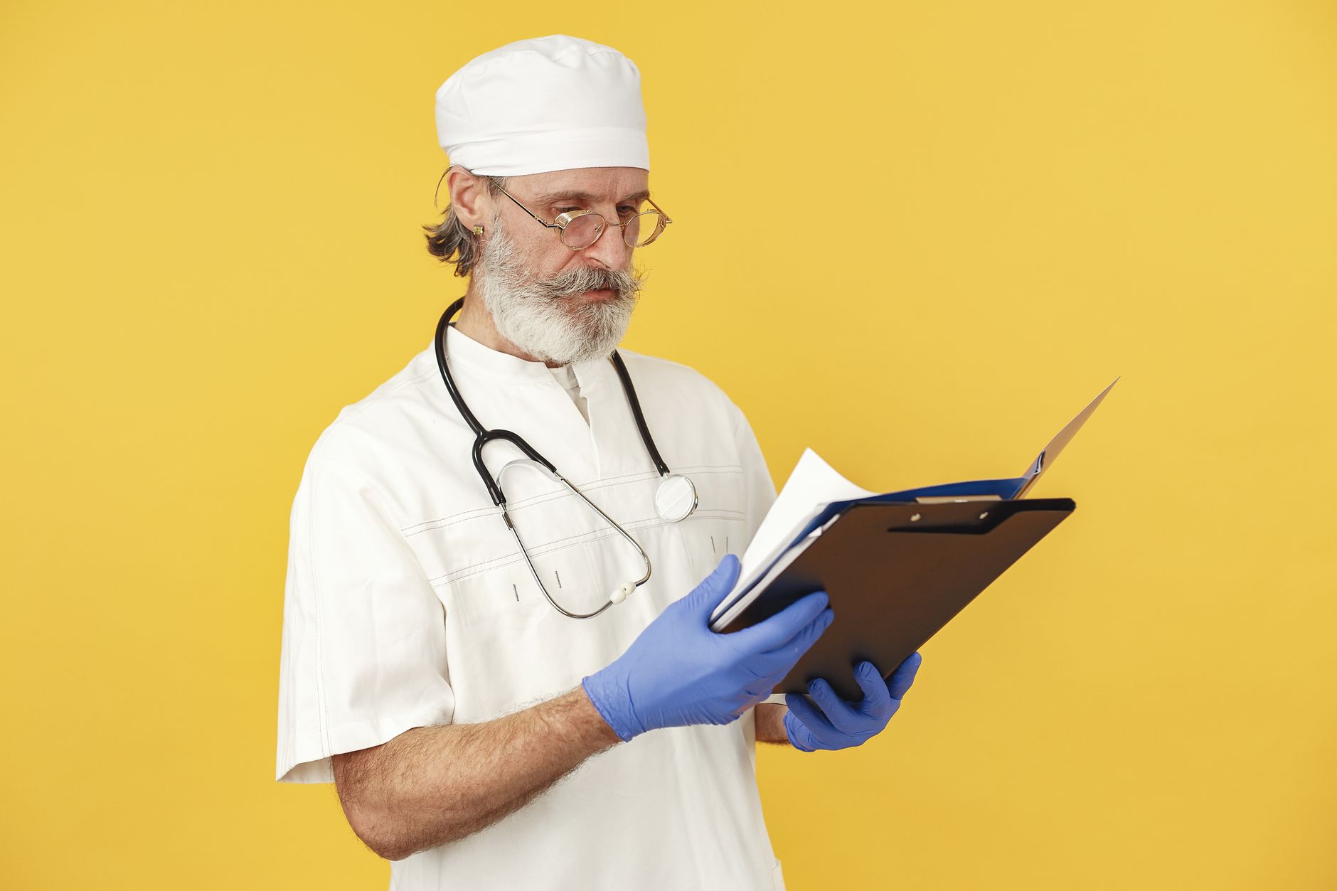 A bearded male doctor with stethoscope reviews medical documents on a yellow background.